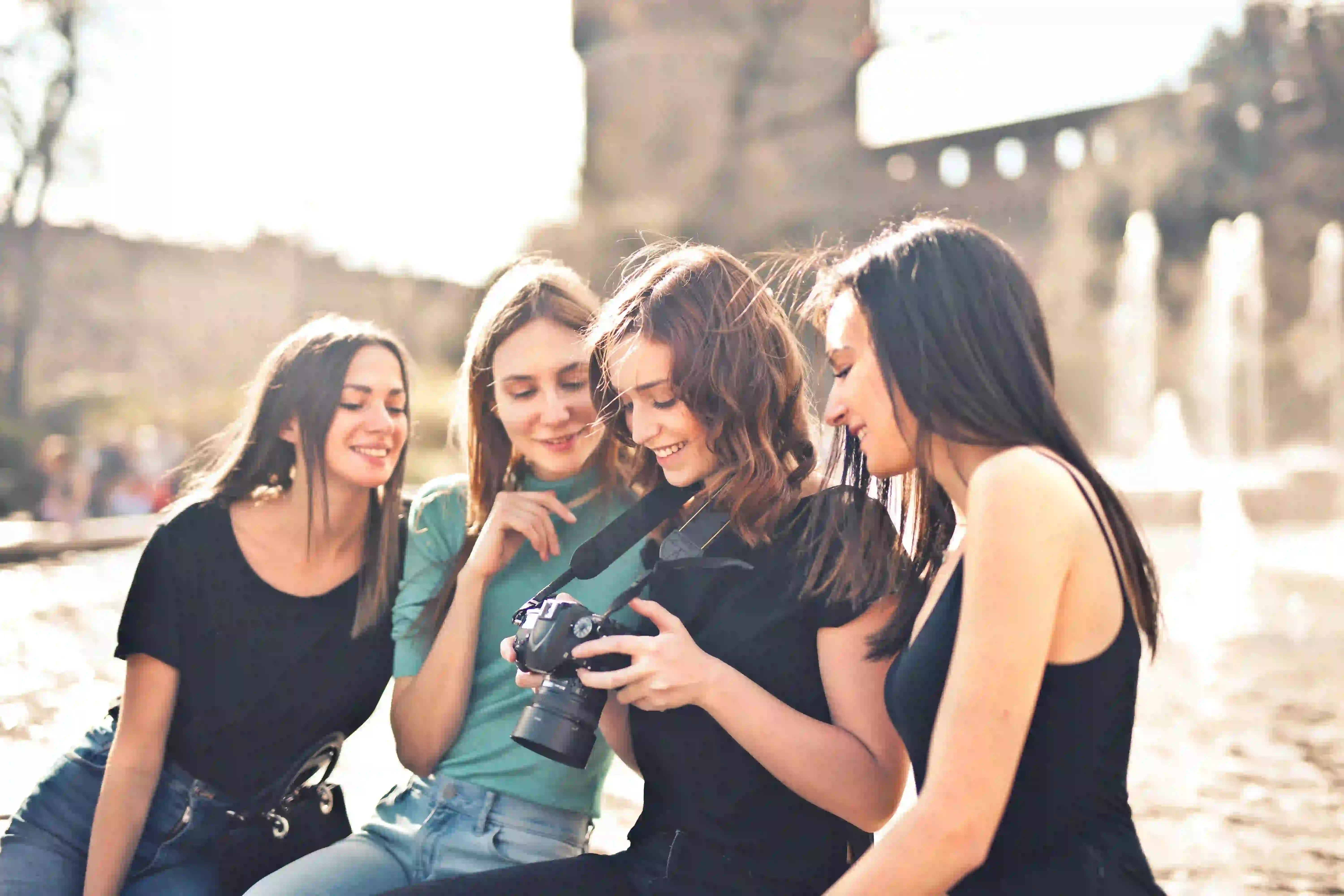 Four young women in distinct outfits are gathered around a camera. They are smiling and interacting, reviewing captured images. They are seated in an outdoor sun-drenched square, with a historical building and fountain in the background. The scene beautifully captures the shared joy of a color walk with friends